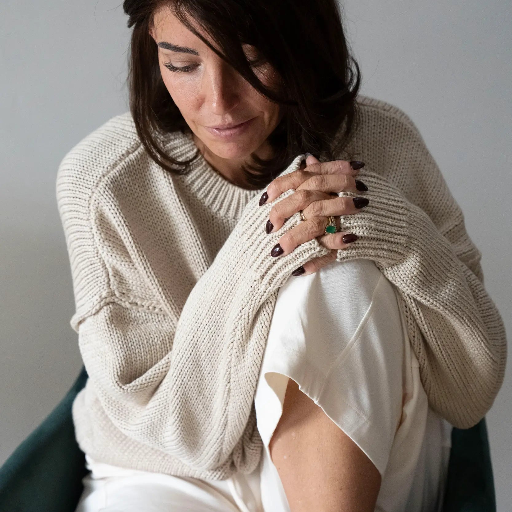 Woman in a cream jumper wearing an Espresso brunette bob wig, sitting in a green chair looking down