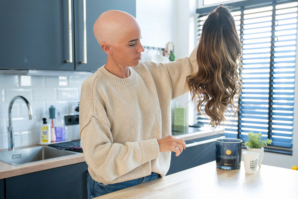 Natalie, She Is Wearing It founder with alopecia, holding up a long wavy brunette human hair wig in her kitchen